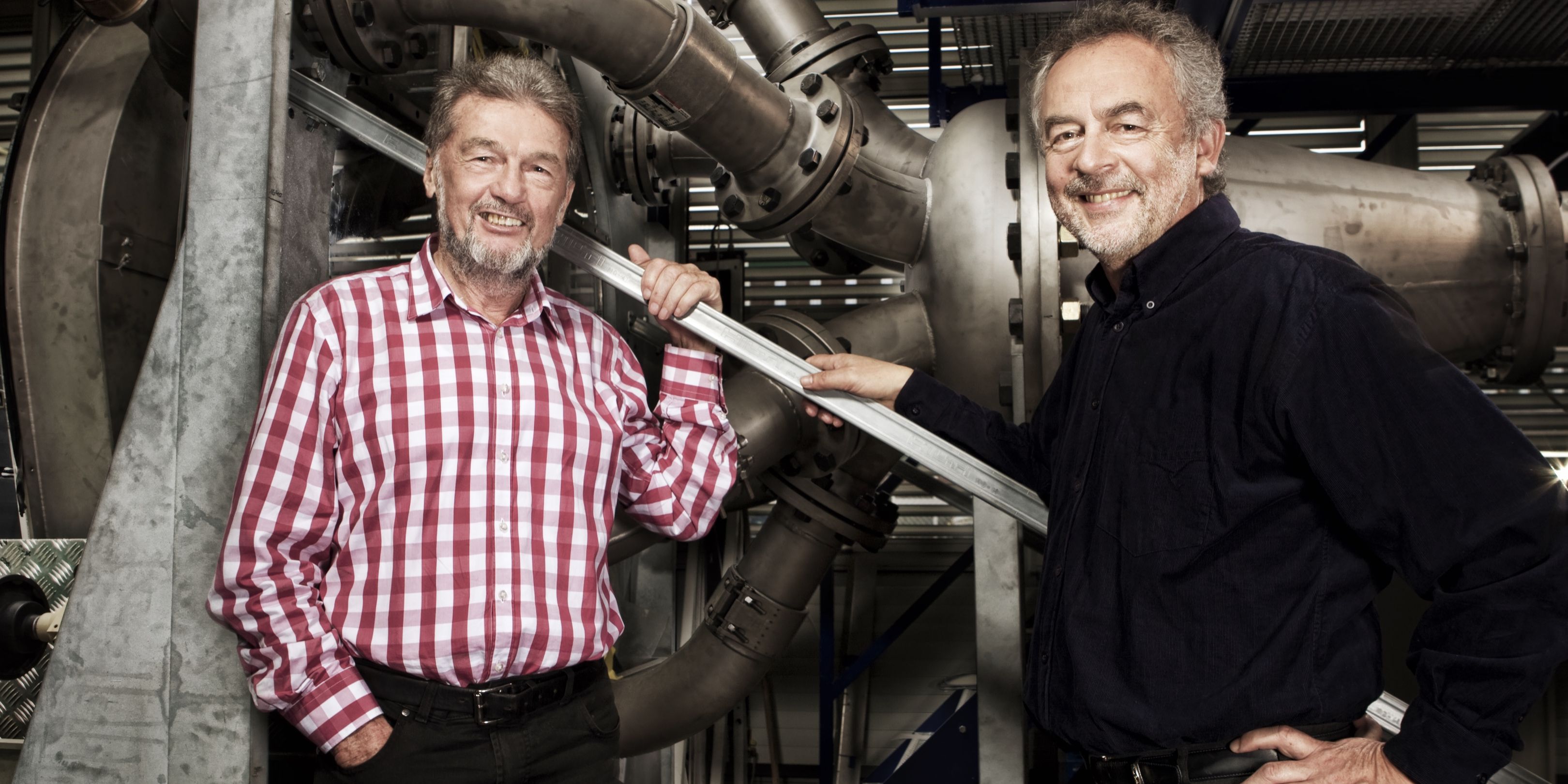 Josef Erlach im karierten Hemd und Prof. Thomas Staubli im schwarzen Hemd stehen am Prüfstand vor einer horizontalen Pelton-Turbine mit sechs Zuleitungen.
