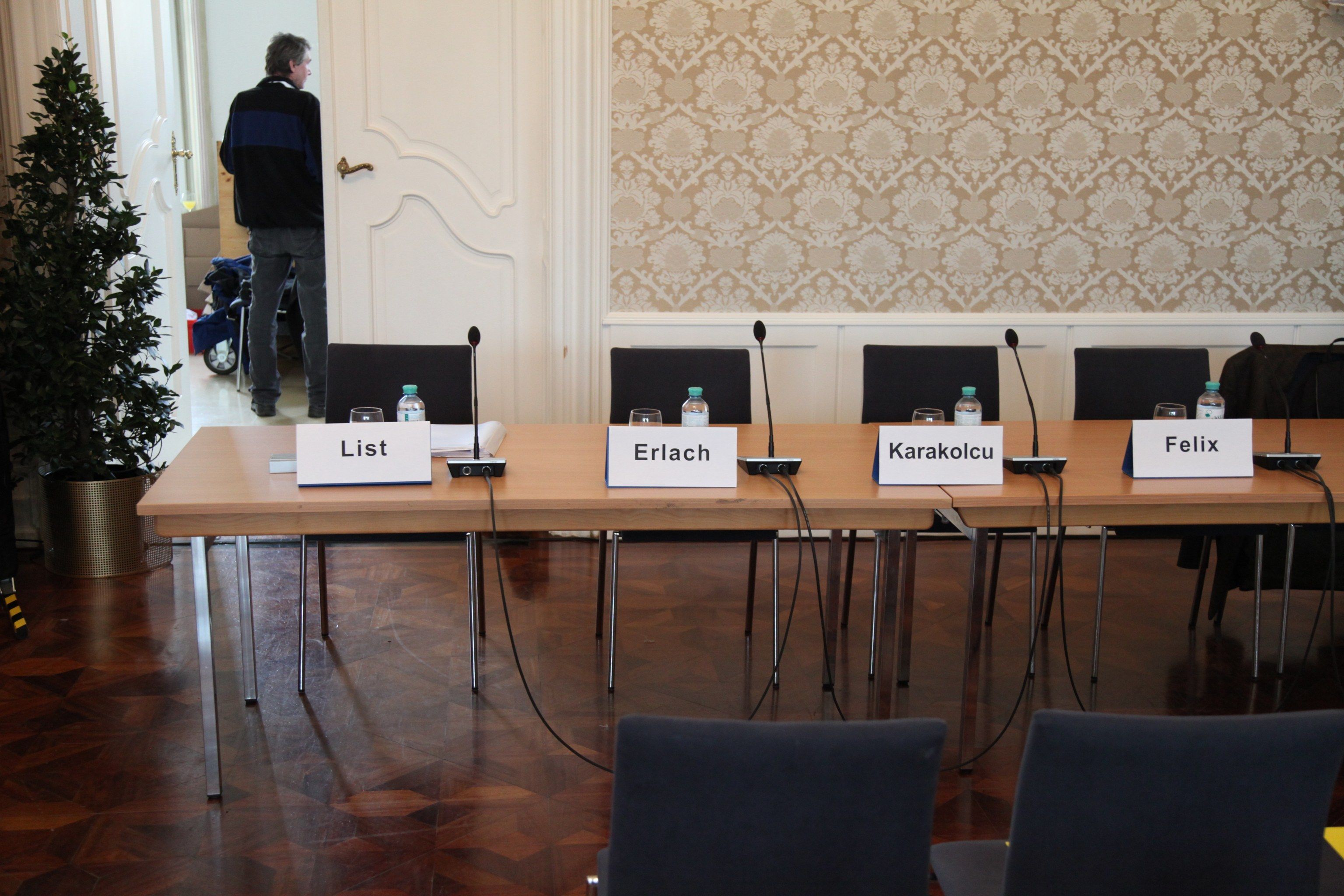 Leeres Podium vor einer gemusterten Tapete, auf dem Tisch vier Namensschilder: List, Erlach, Karakolcu, Felix.