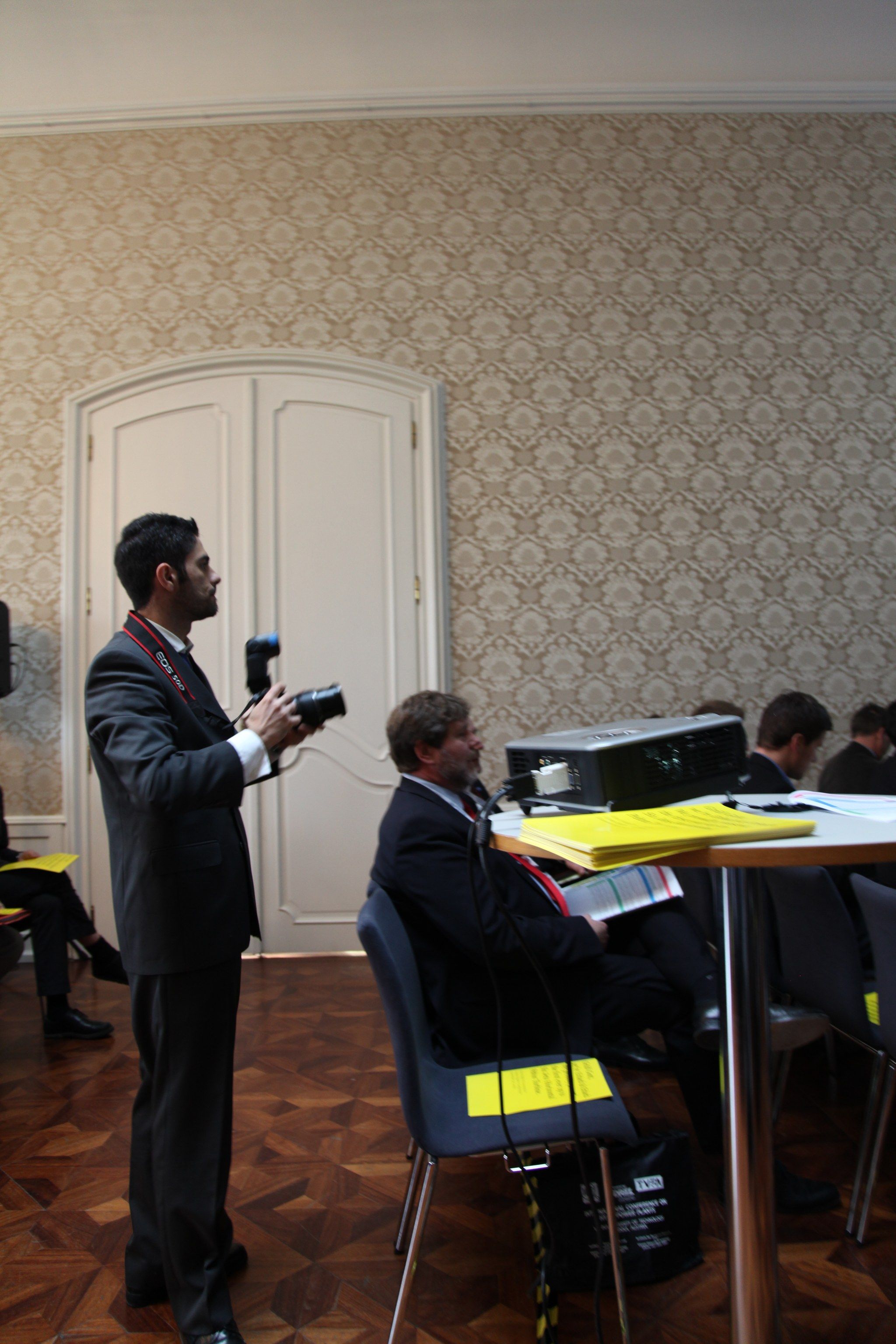 Ein Fotograf steht mit der Kamera im Saal, daneben sitzen Teilnehmer an einem Tisch mit gelben Faltblättern.